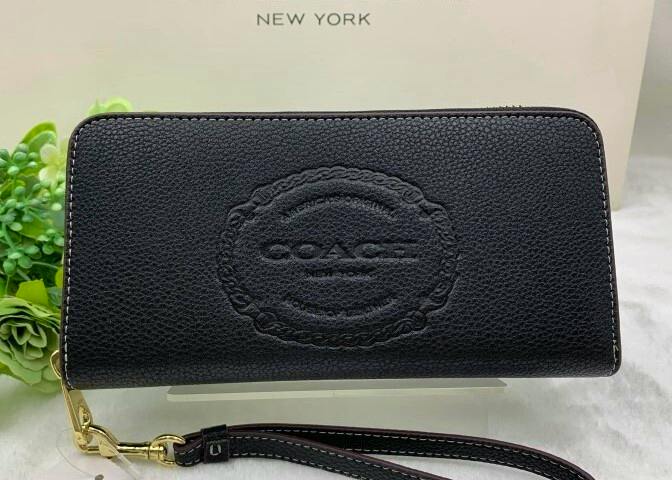 Black COACH long zip wallet in leather with an embossed logo on the front and a wrist strap, shown on a light background.