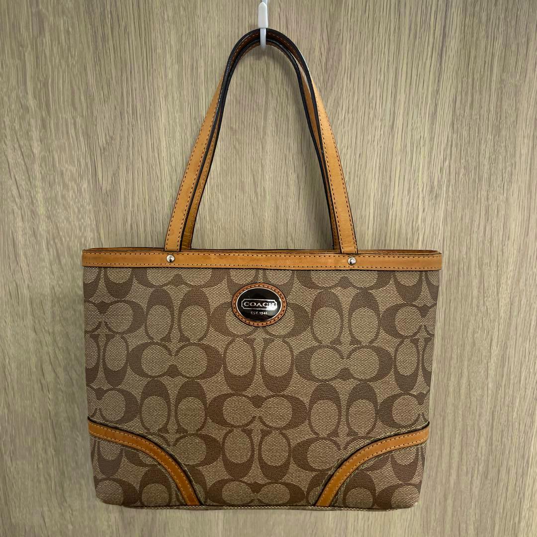 Front view of a COACH mini tote bag in brown Heritage Signature canvas hanging on a hook against a wooden wall.