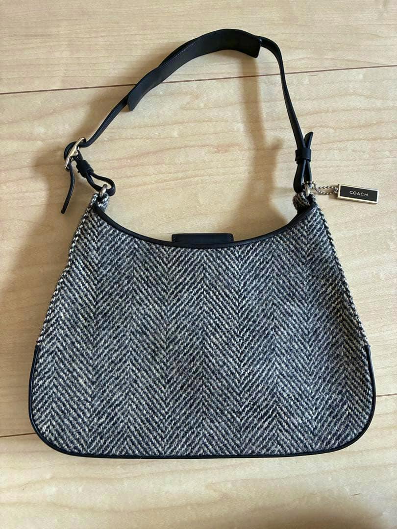 Front view of a Coach herringbone canvas handbag with black leather trim and shoulder strap laid flat on a wooden floor.