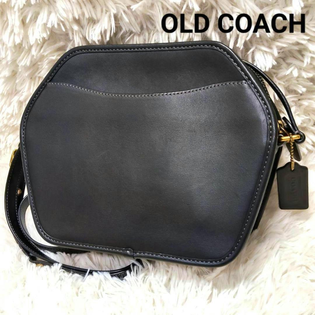 Front view of a black hexagonal Coach leather shoulder bag on a white shag background with a leather hang tag and shoulder strap visible.