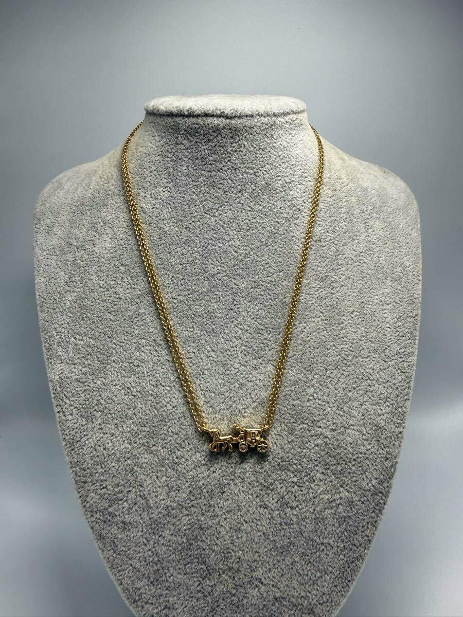 Gold-colored necklace with a Coach horse-and-carriage pendant displayed on a gray jewelry bust.