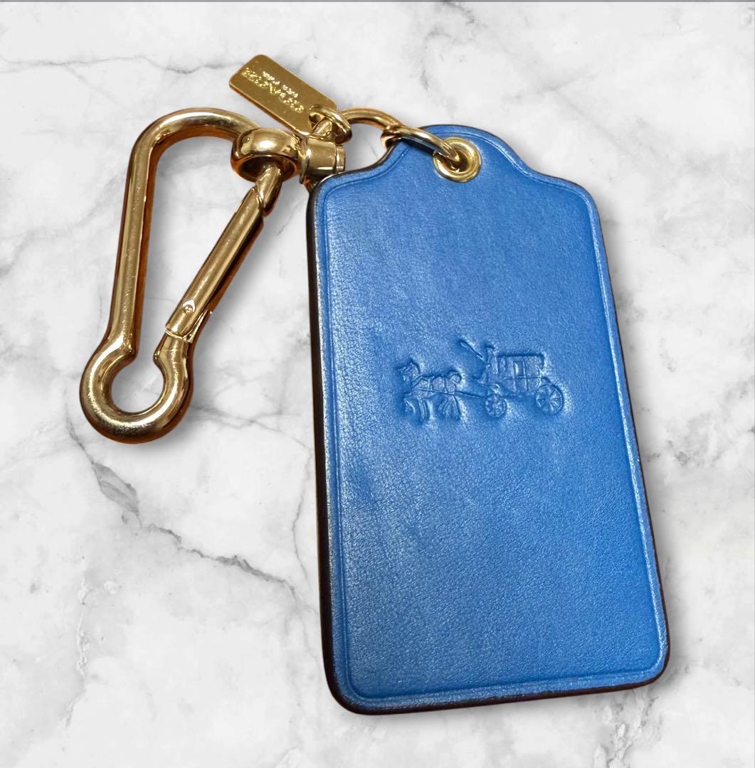 Blue leather COACH horse-and-carriage bag charm keychain with gold hardware on a marble background.