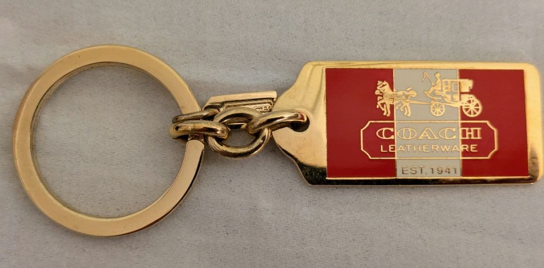 A zinc-alloy keyring with a rectangular metal plate showing the Coach horse-and-carriage logo on a gray background.
