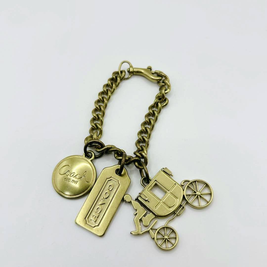 Brass-tone COACH horse-and-carriage bag charm key fob in zinc alloy, showing the chain with a carriage pendant and two COACH tag charms.
