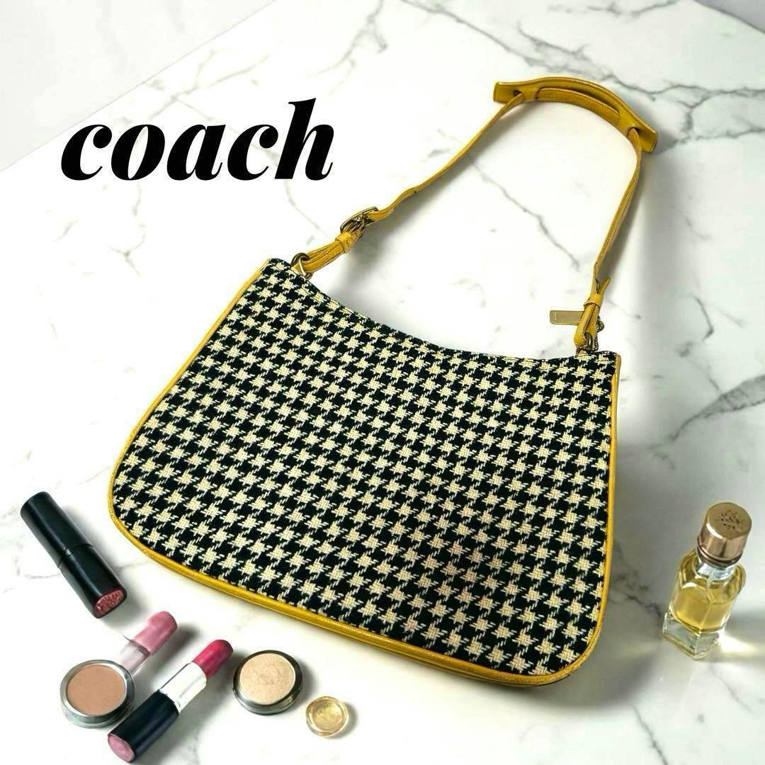 A houndstooth-patterned Coach shoulder bag with yellow leather trim displayed on a marble surface alongside makeup items.