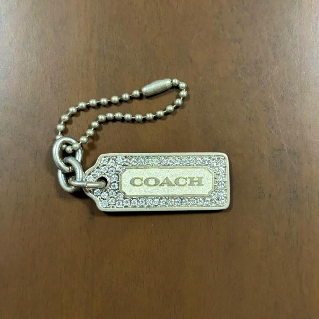 Metal Coach bag charm keychain tag with COACH logo and crystal border on a brown surface.