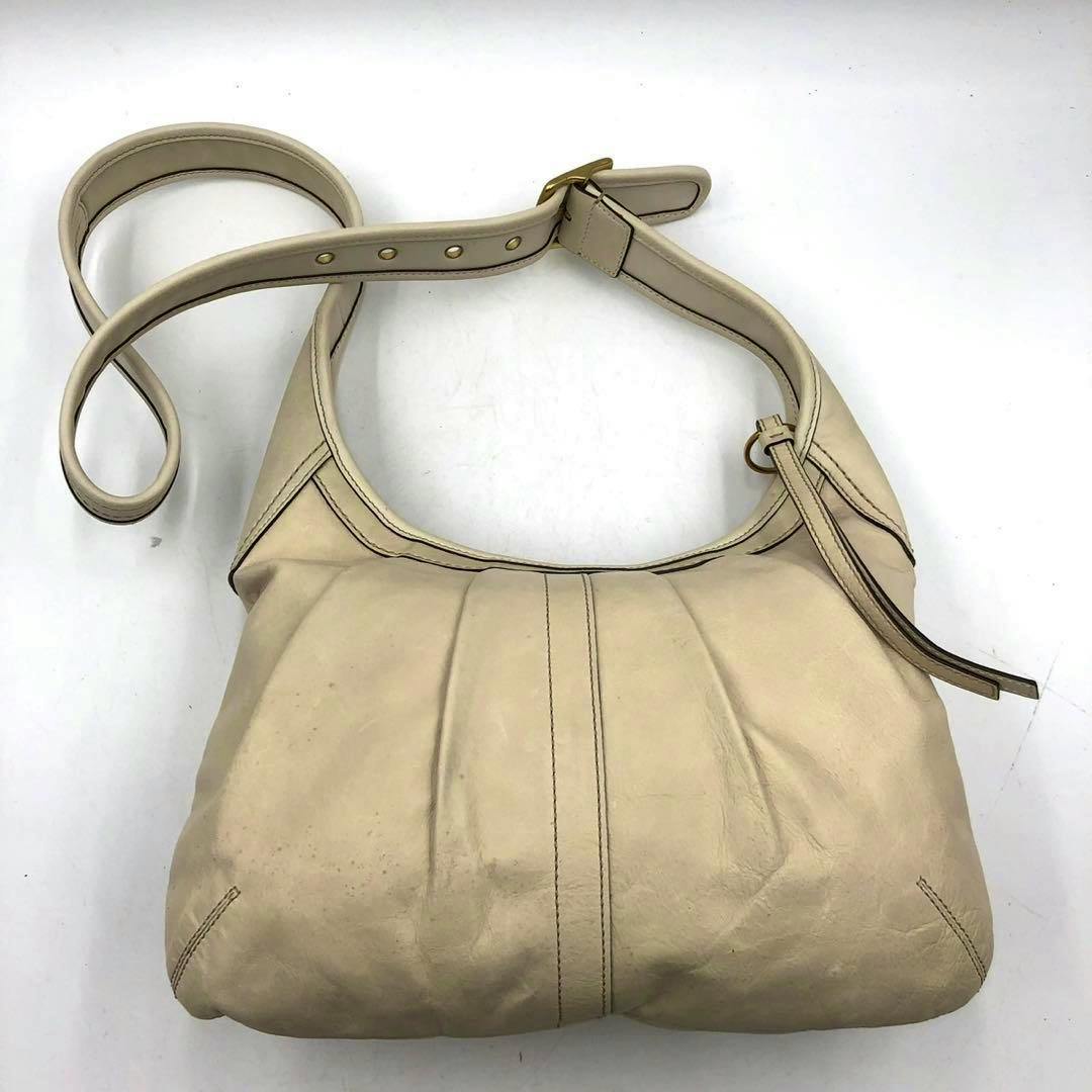 Ivory-colored Coach shoulder bag made of synthetic leather shown flat with its strap and front pleat details visible.