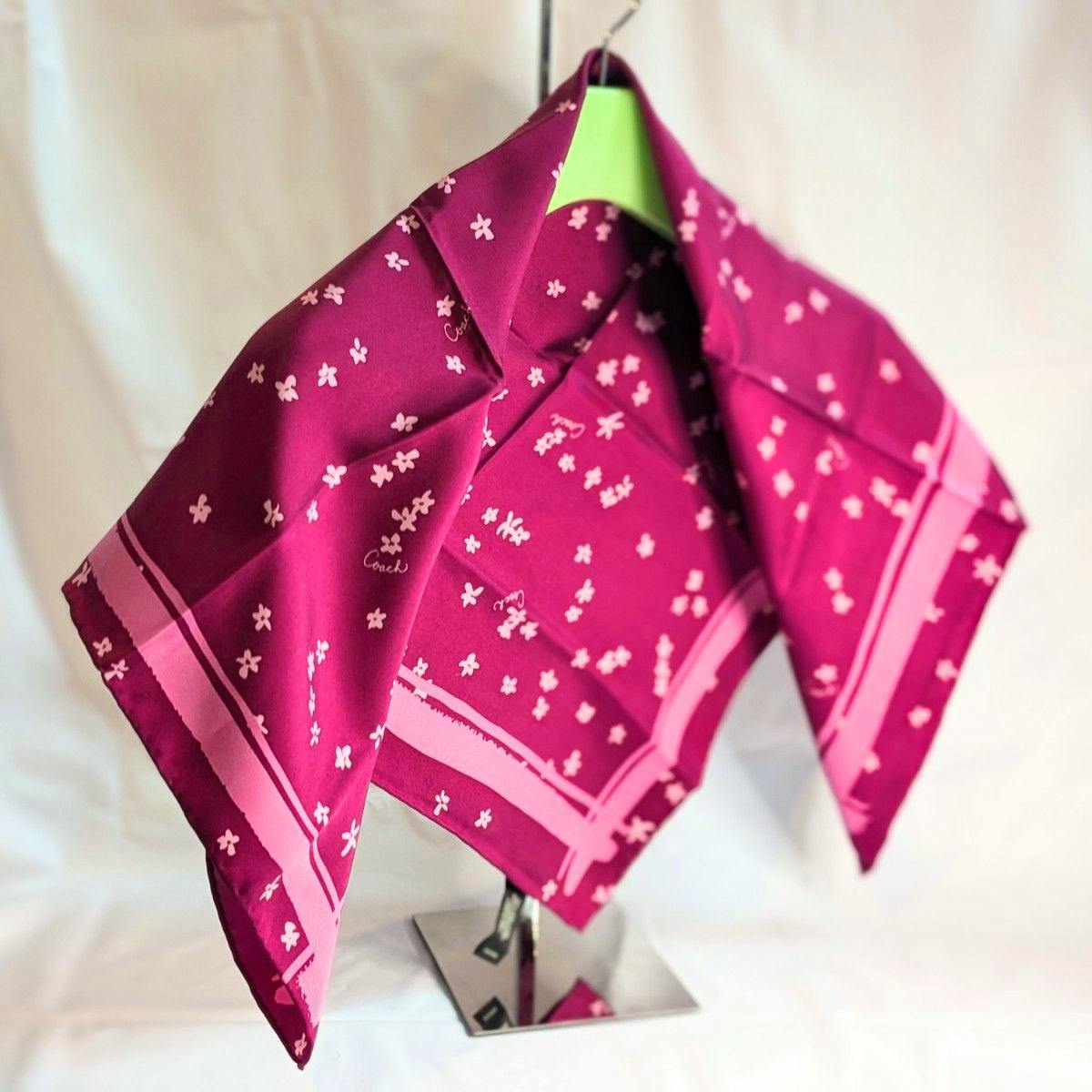 Large pink Coach silk scarf with small floral print and light-pink border draped on a display stand.