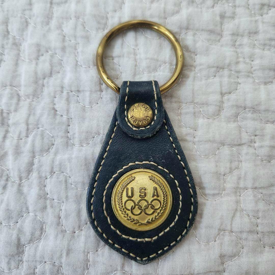 Black leather Coach keyfob with a brass ring and a gold-tone circular USA Olympic medallion centered on the fob.