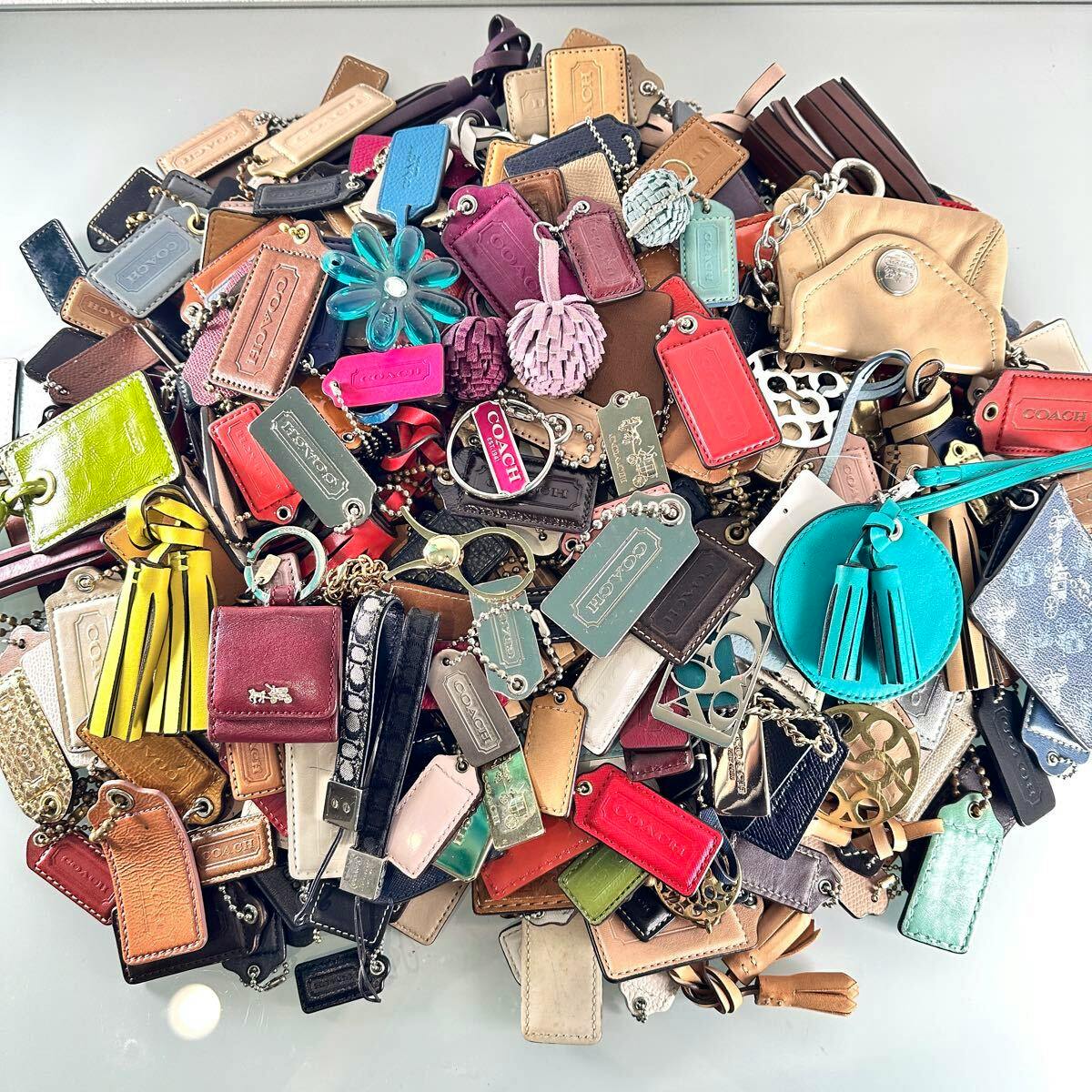 Pile of assorted COACH leather keychain tags, charms, tassels, and metal keyrings on a reflective surface.