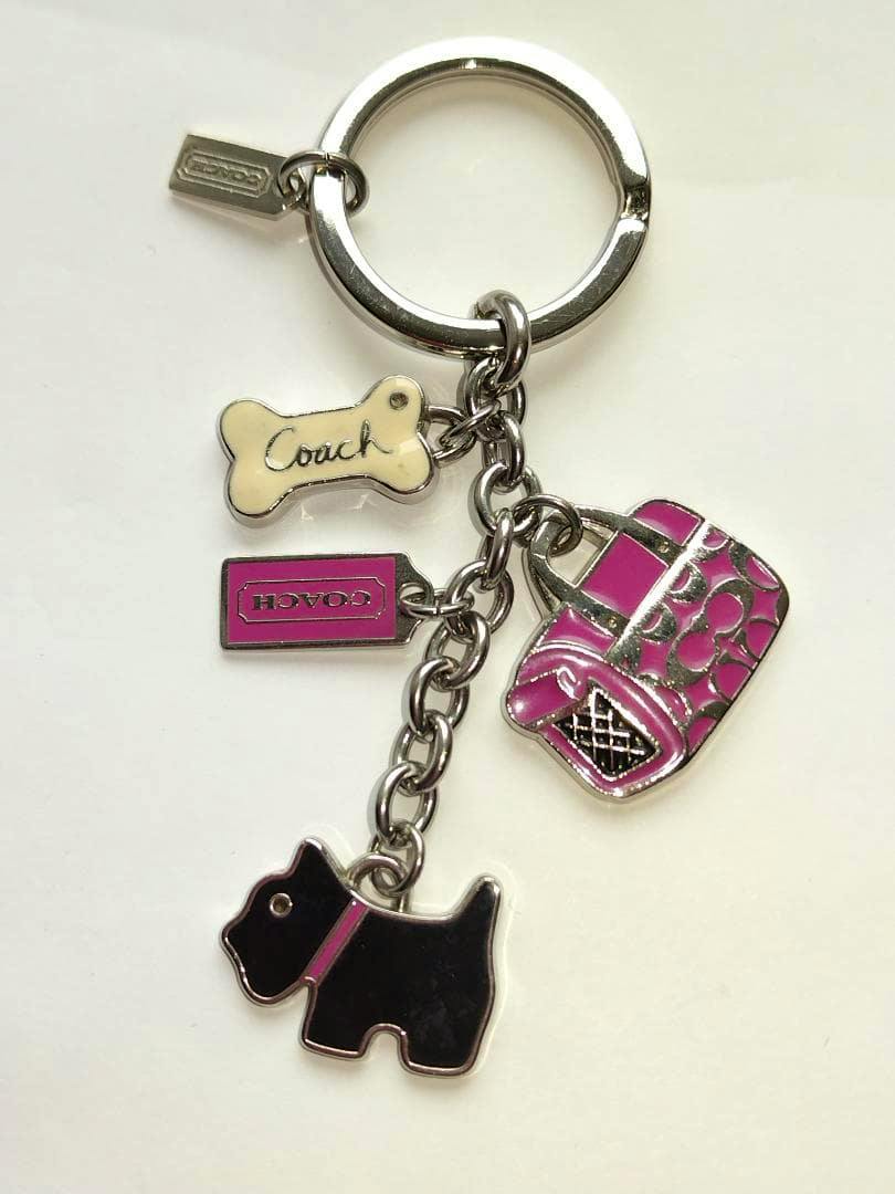 Front view of the COACH keychain showing a metal keyring with attached enamel charms: a black dog, a pink handbag, a bone, and a rectangular tag.
