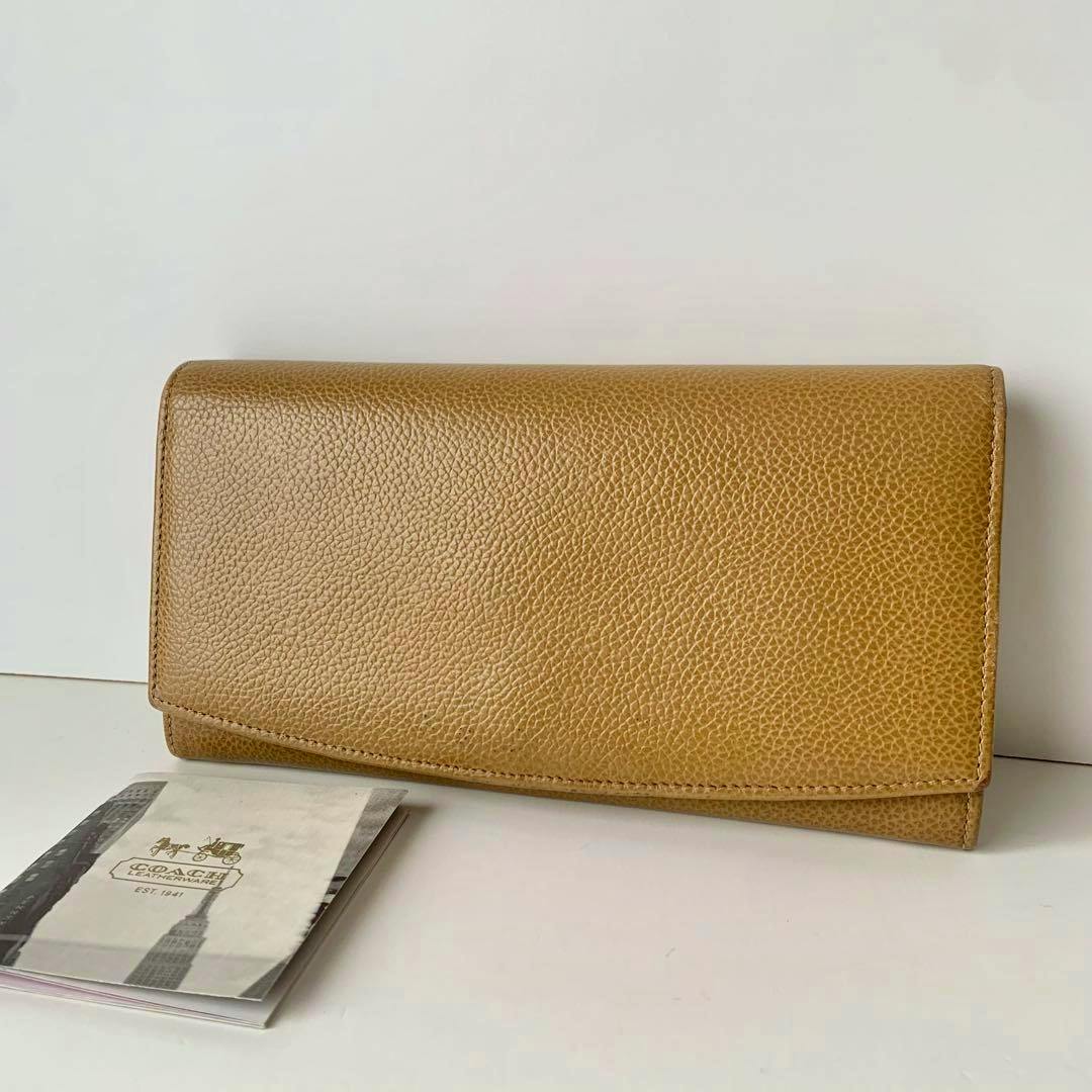 Tan pebbled-leather Coach long wallet (closed flap) displayed on a white background with a small Coach booklet beside it.