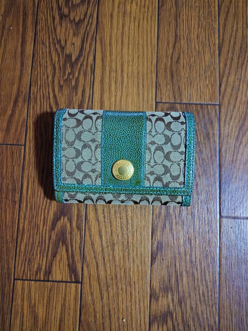 A closed COACH L-shaped zipper canvas wallet with signature C pattern and green trim placed on a wooden floor.
