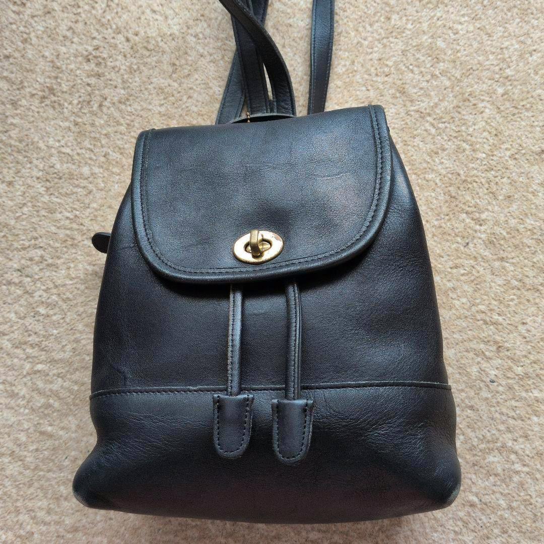 Front view of a black COACH leather backpack with a flap, turn-lock closure, drawstring straps, and shoulder straps visible.