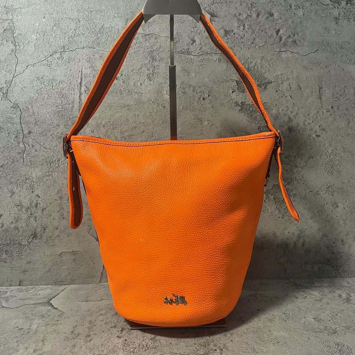 Front view of an orange leather Coach bucket shoulder bag displayed on a stand against a gray background.