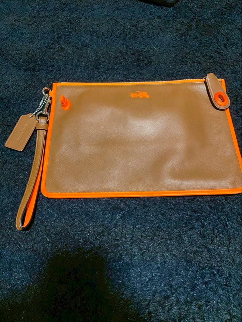 Beige-orange leather Coach clutch bag with orange trim and wrist strap on a dark textured background.