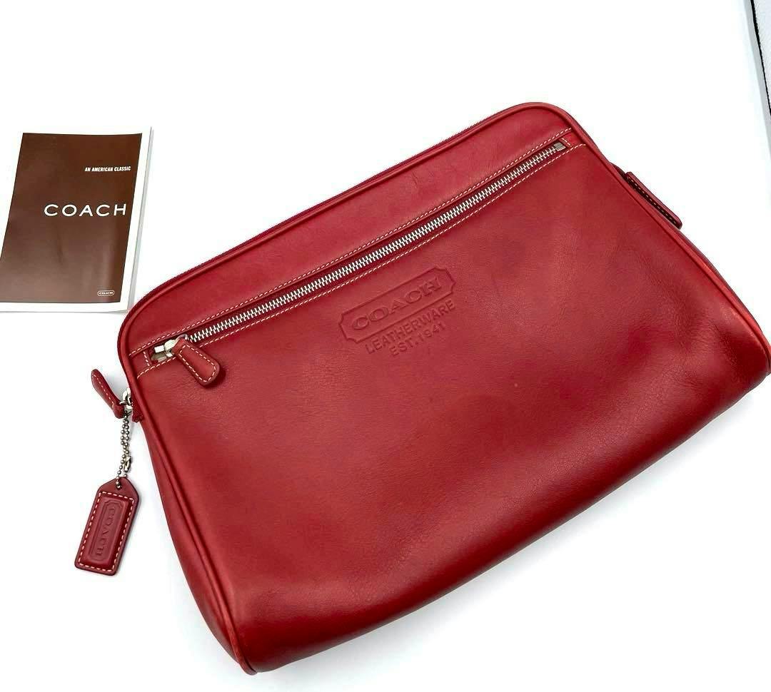 Red Coach leather clutch bag with a front zip pocket and attached leather tag on a white surface with a Coach booklet nearby.