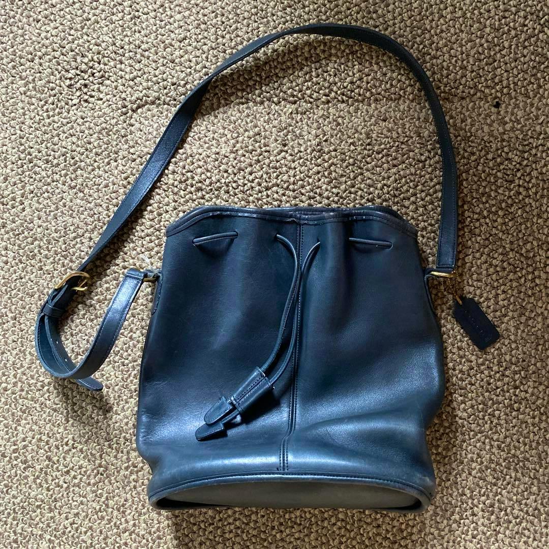 Black COACH leather drawstring (bucket) shoulder bag lying flat on a woven carpet, showing the strap and drawstring closure.