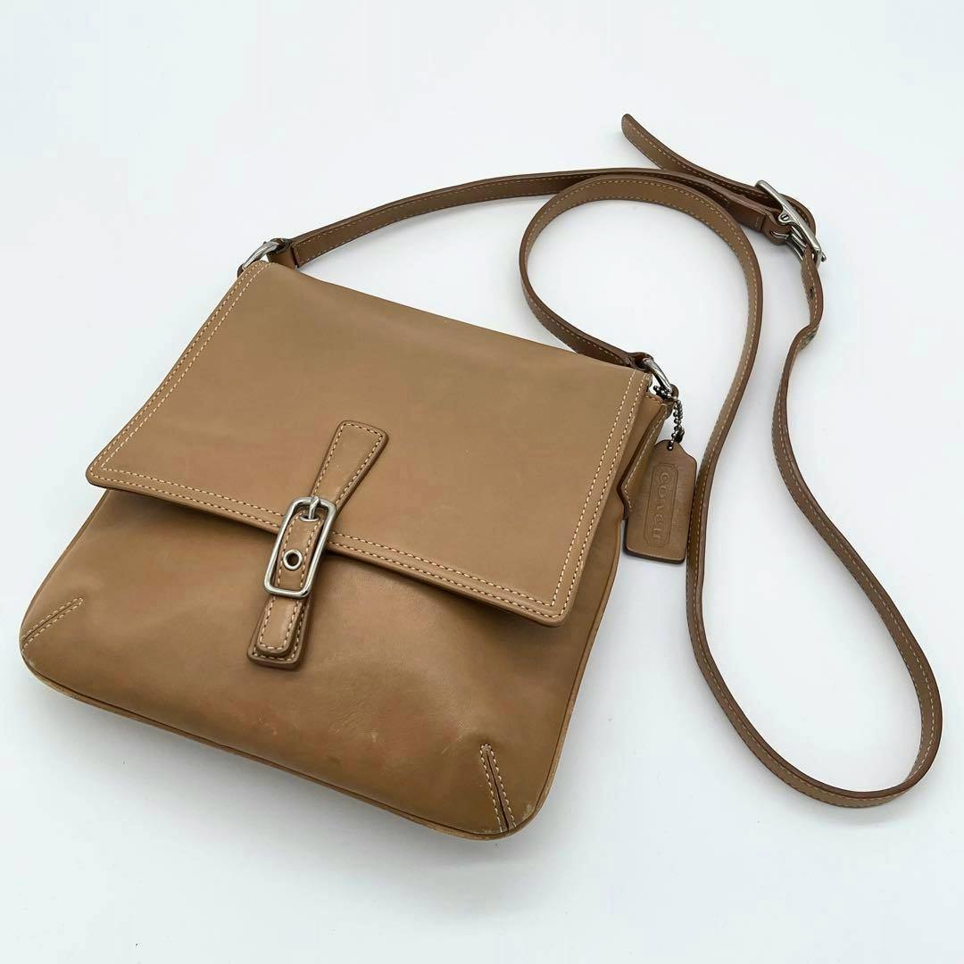Light brown Coach leather flap shoulder bag with front buckle and long adjustable strap laid flat on a white background.