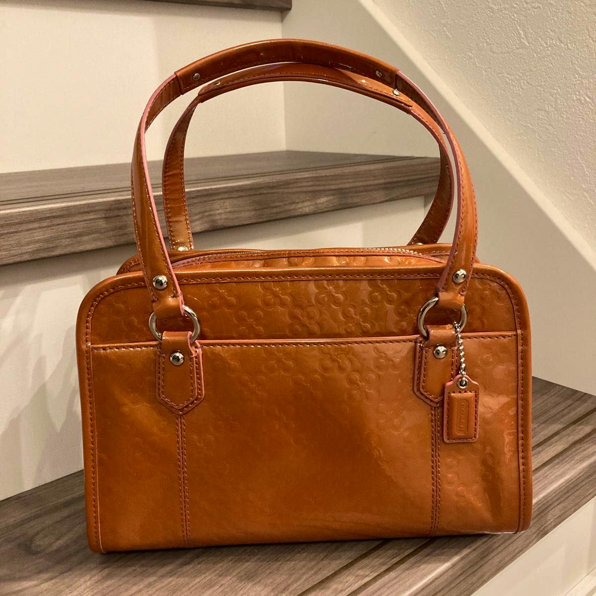 Front view of an orange COACH cow leather handbag with double handles and a small leather tag, photographed on wooden stairs.
