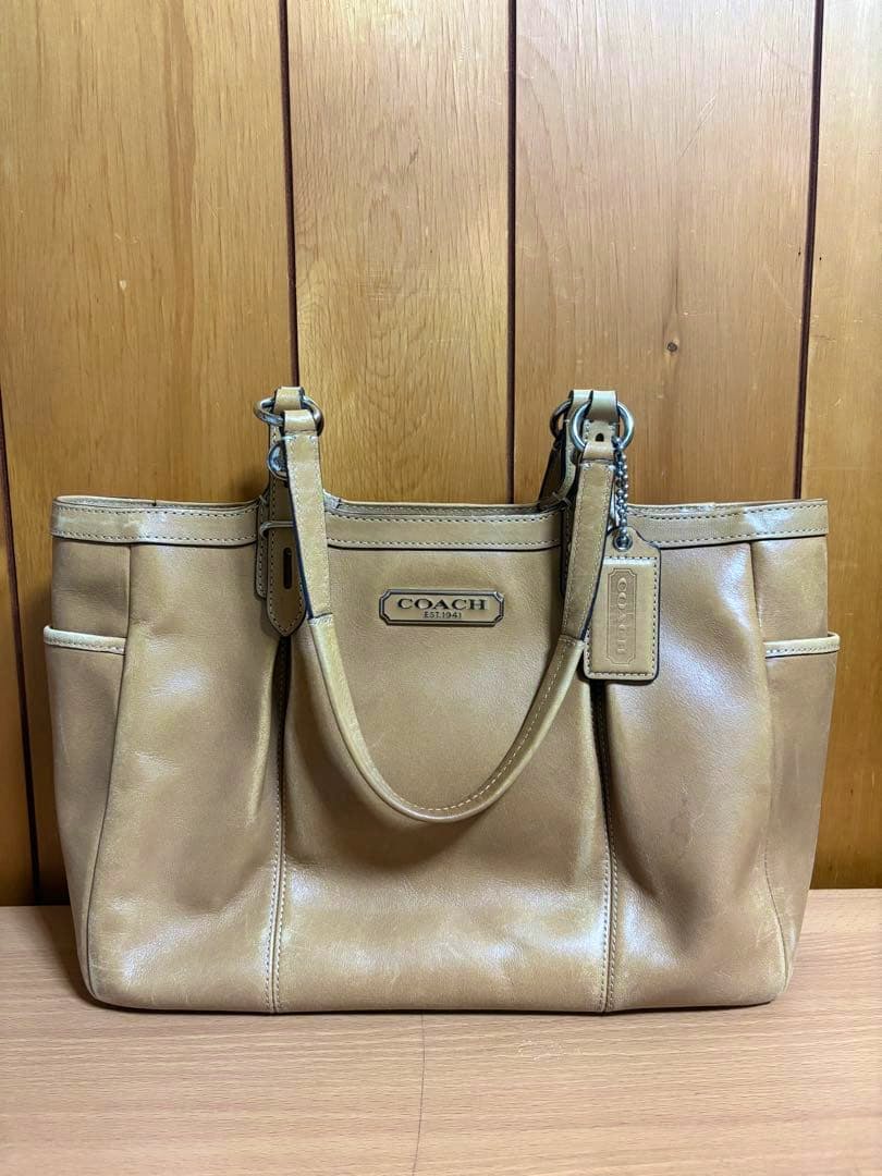 Front view of a beige COACH leather handbag with two handles and a hanging COACH tag against a wooden background.
