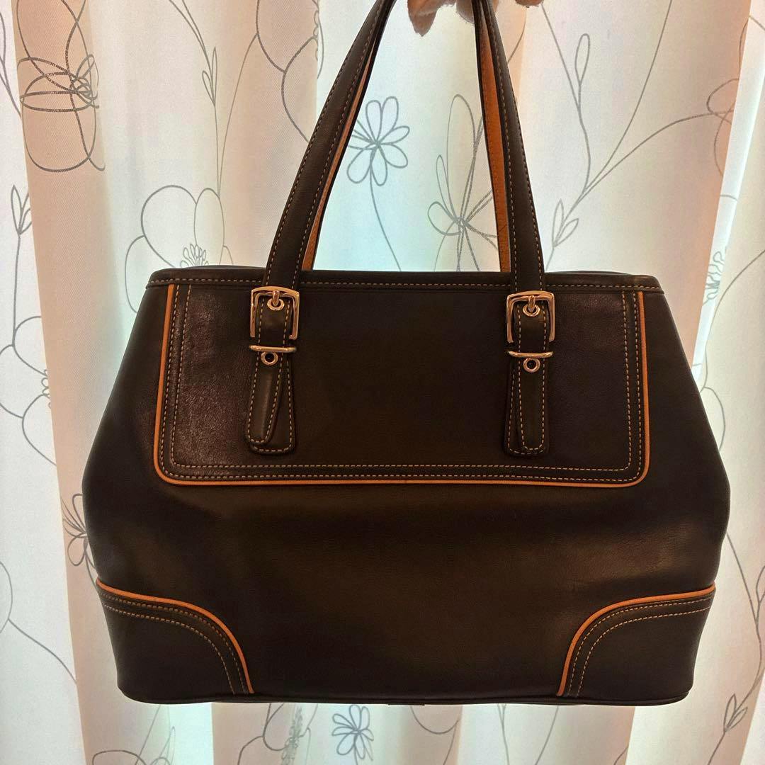 Dark brown cow leather Coach handbag with double top handles, contrasting trim, and buckle accents photographed against a light curtain background.