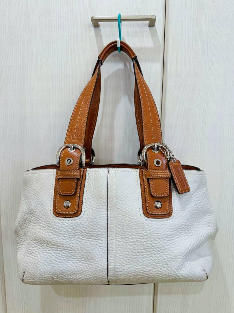 White and brown leather COACH handbag hanging on a hook against a light-colored wall.