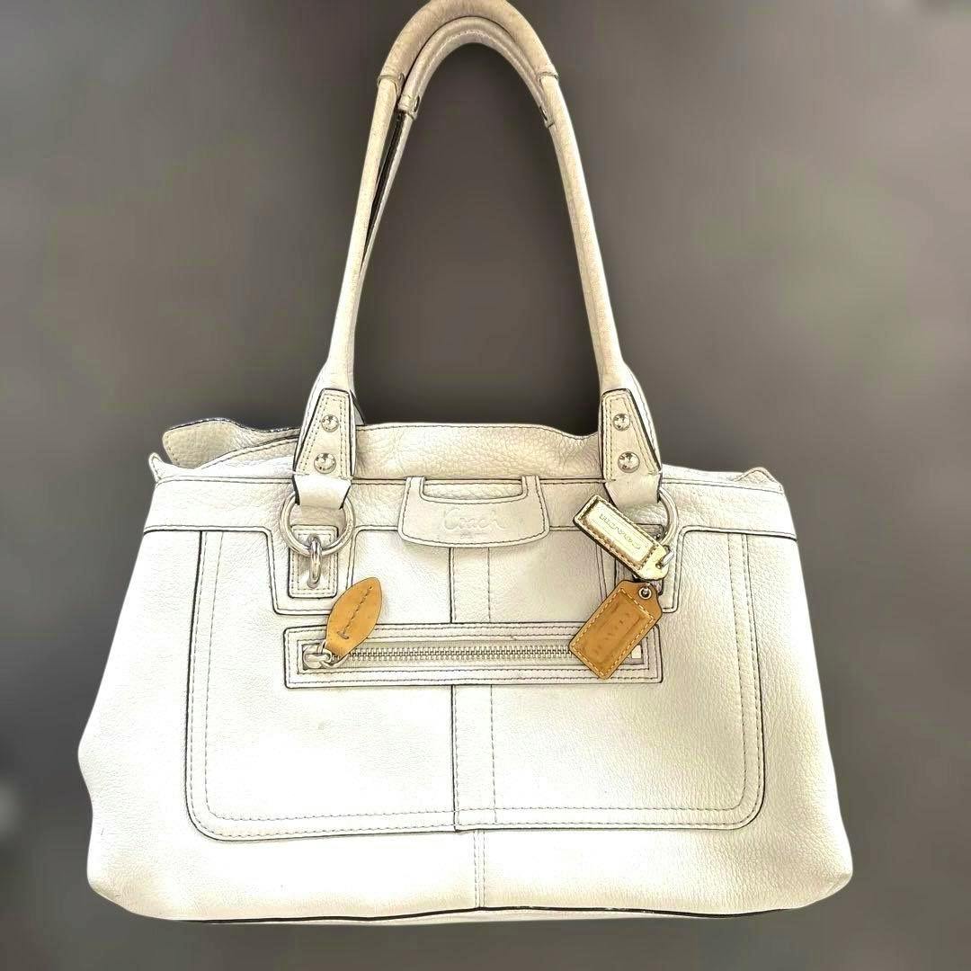 Front view of a white Coach leather handbag with double handles, a front zip pocket, and attached leather tags.