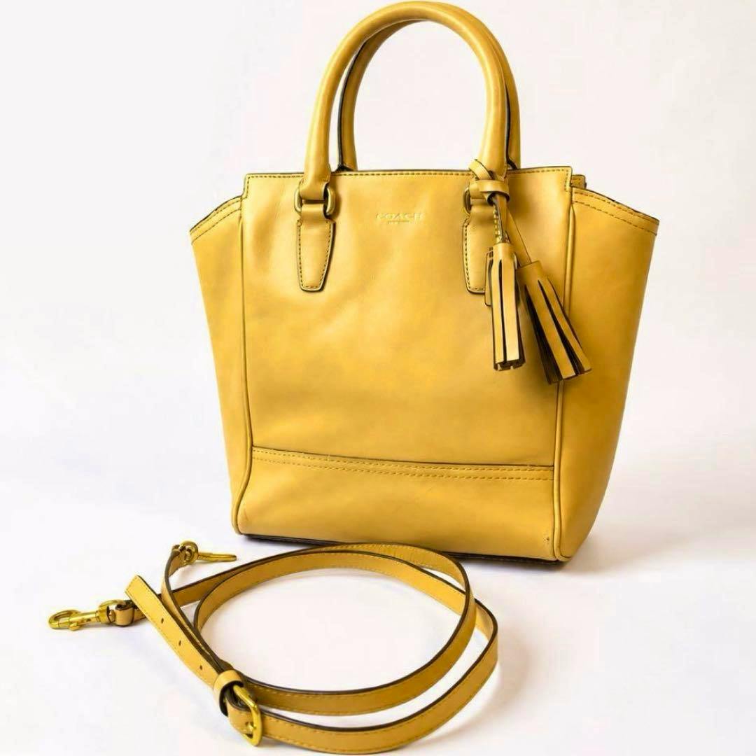 Yellow COACH leather handbag with top handles and tassel, shown with a coiled detachable shoulder strap on a white background.