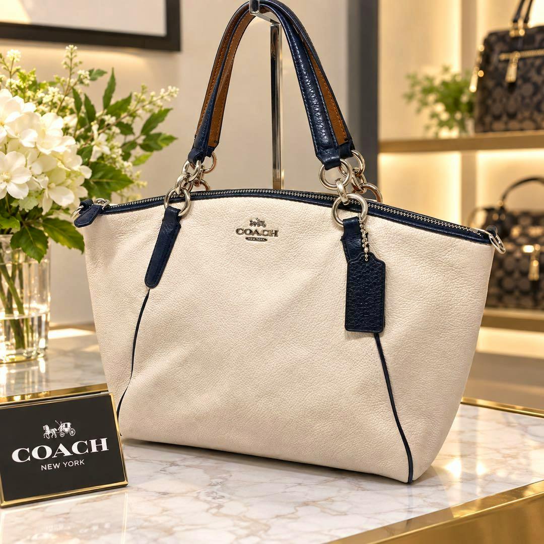 Cream leather COACH handbag with navy trim and double handles displayed on a marble countertop.