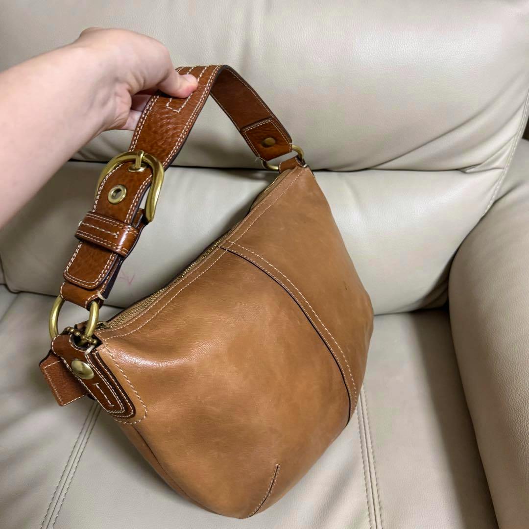 COACH brown leather hobo shoulder bag with a wide buckle strap being held by a hand against a light leather chair.