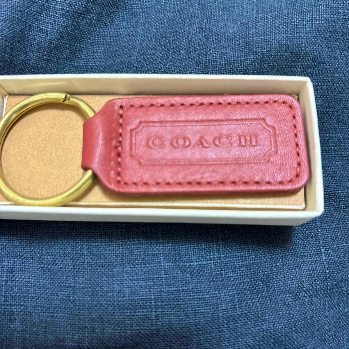 Red leather COACH keyring with a gold metal ring displayed in a small box on dark fabric.