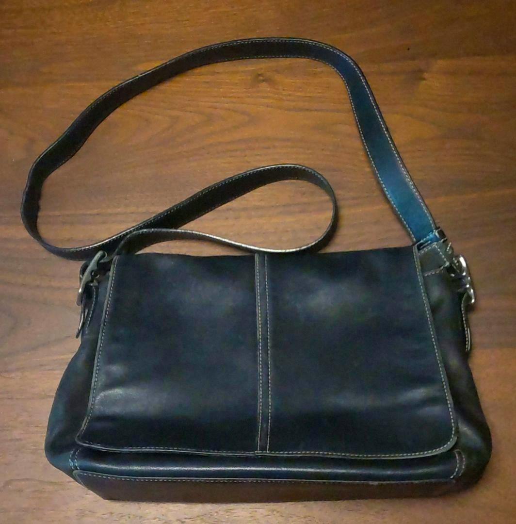 Black leather Coach shoulder messenger bag with flap and long strap laid flat on a wooden surface.