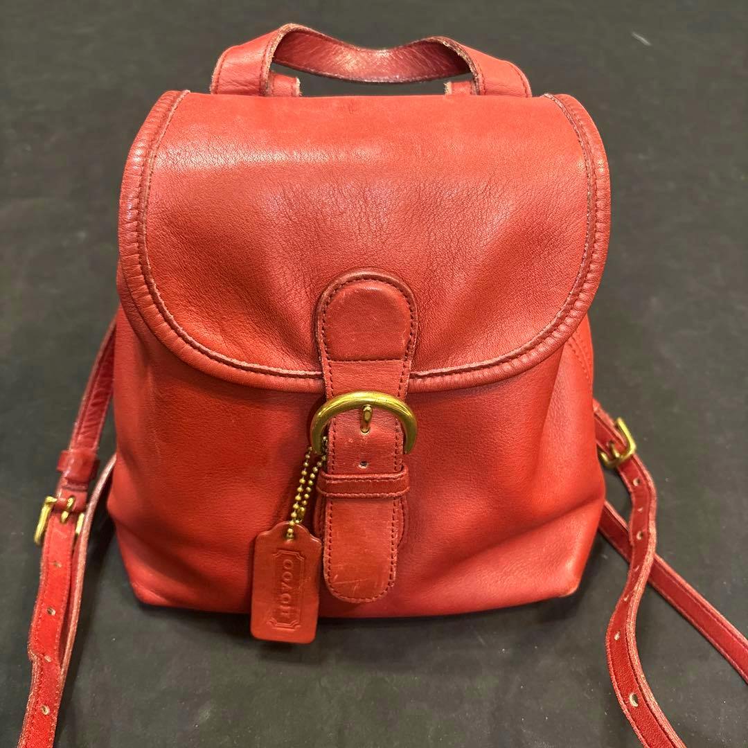 Front view of a red leather mini backpack with a flap, buckle, and Coach hang tag.