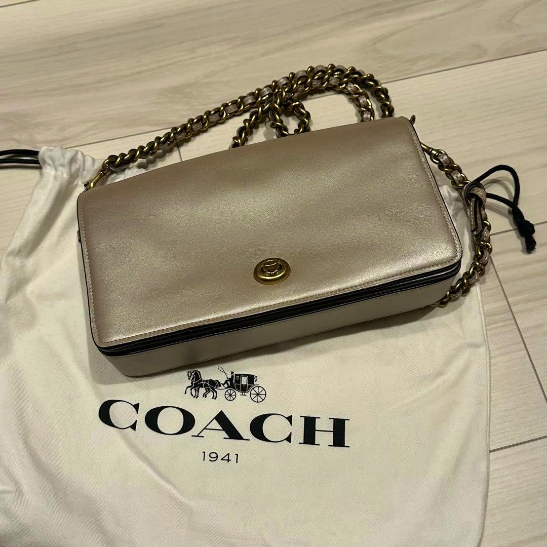 Light beige Coach leather shoulder bag with a gold chain strap displayed on a white Coach dust bag.