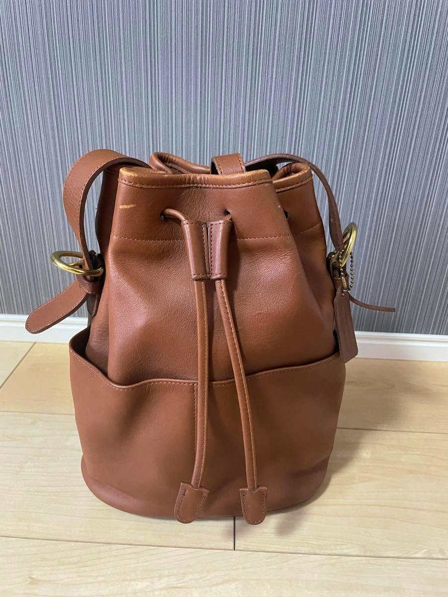 Brown leather Coach drawstring shoulder bag standing on a light wood floor against a striped wall.