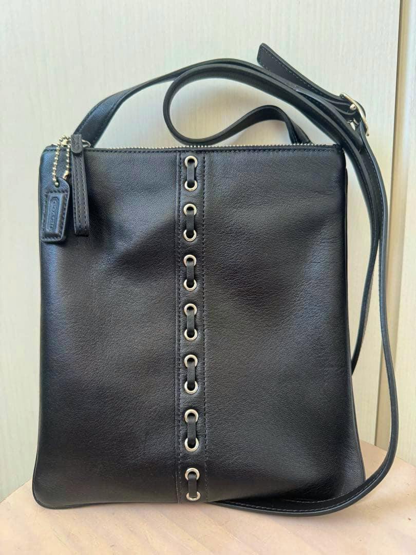 Front view of the black COACH leather shoulder bag showing a vertical row of metal grommets, leather strap, and hang tag.