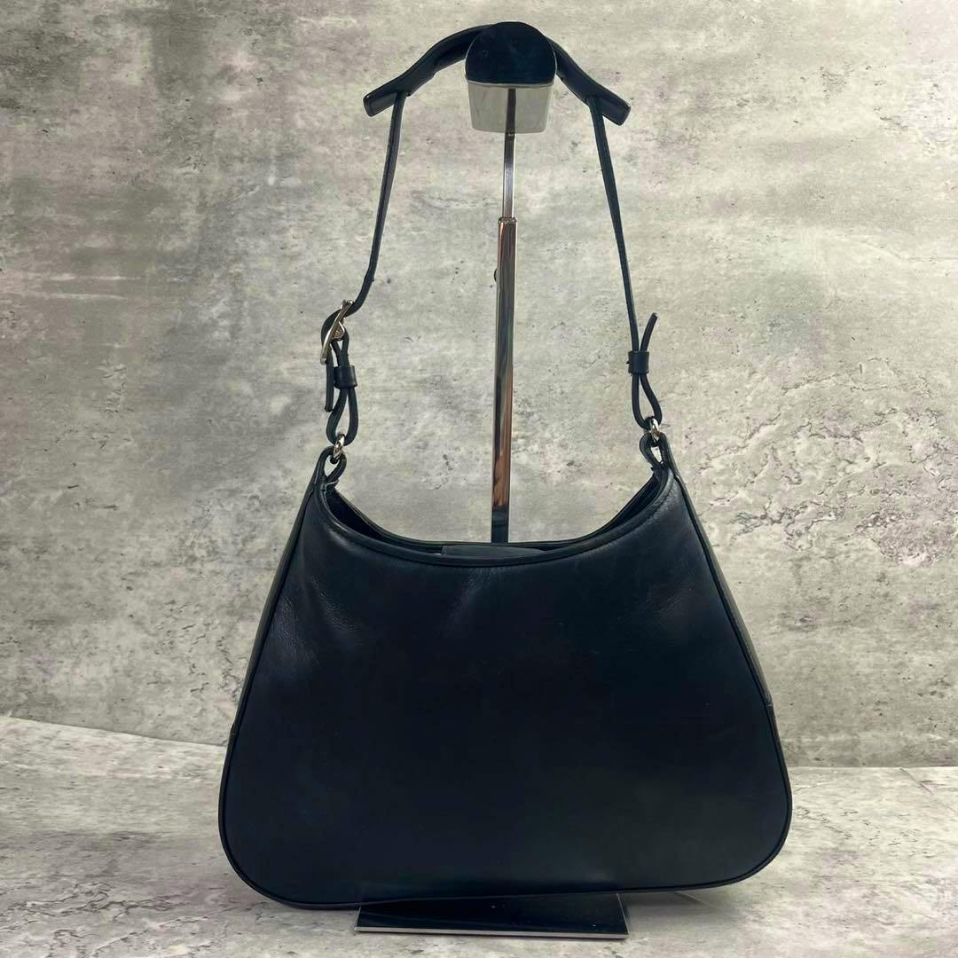 Black leather Coach one-shoulder handbag hanging on a display stand against a gray background.