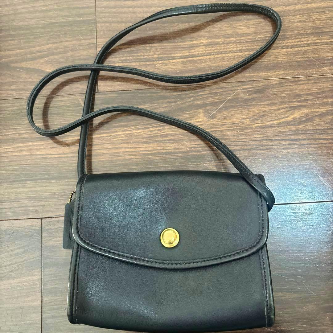 Front view of a black Coach leather shoulder bag with a flap closure and a round gold snap, shown with its long strap on a wooden floor.