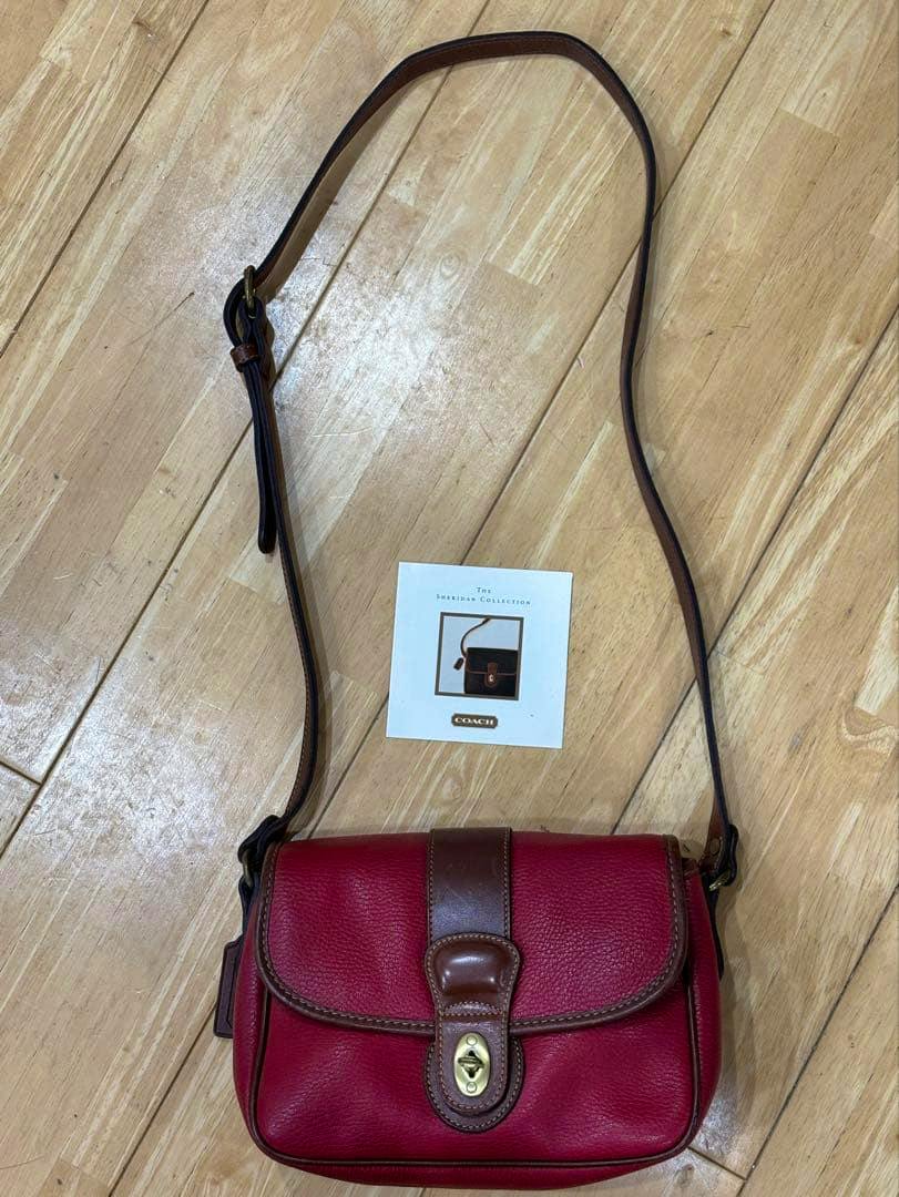 Front view of a red COACH leather shoulder bag with brown trim, gold turn-lock, and an adjustable strap placed on a wooden floor with a small Coach card nearby.