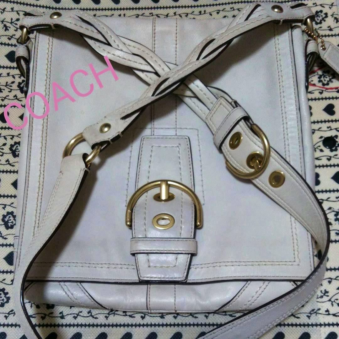 A Coach leather shoulder bag with a gold-tone buckle and crossed shoulder straps laid flat on patterned fabric.