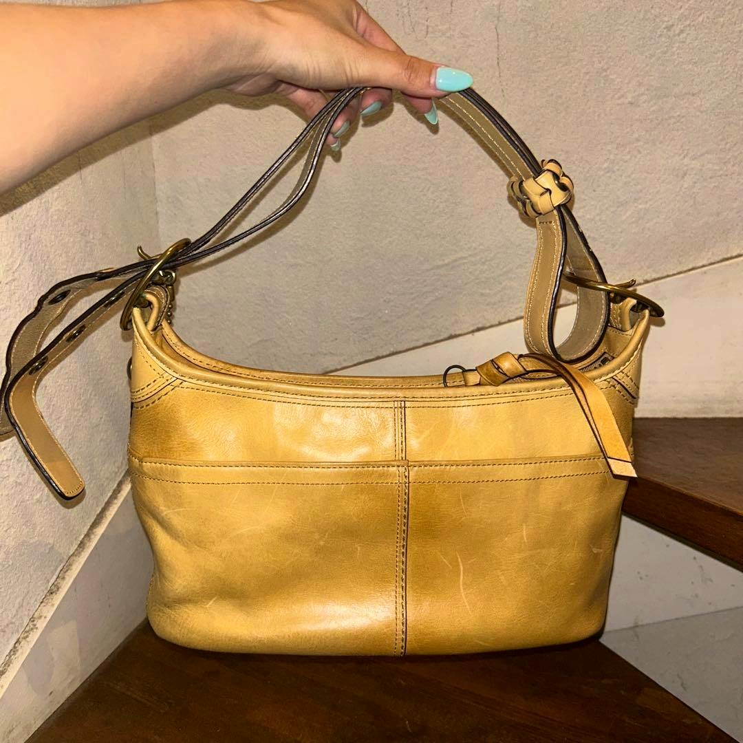 Tan COACH cow-leather shoulder and hand bag shown front-on with its strap held by a hand.