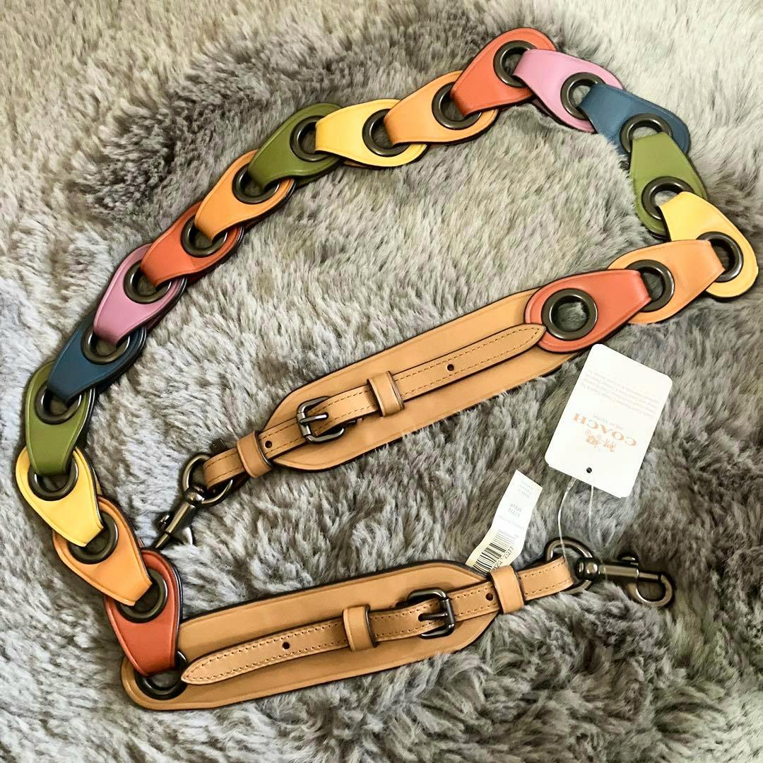 A multicolored Coach leather shoulder strap with buckles, metal clasps, and attached tags laid on a gray fur surface.