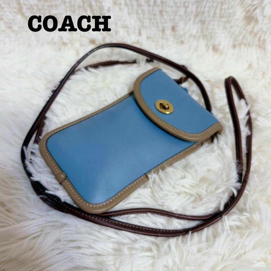 Blue Coach leather smartphone shoulder bag with tan trim, turnlock closure, and brown strap lying on a white fur background.