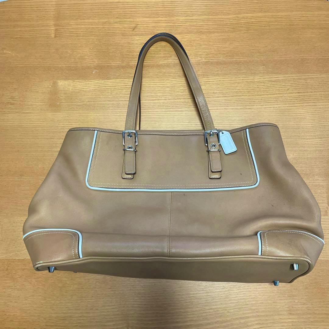 Beige COACH leather tote bag laid flat on a wooden surface, showing the front, straps, and hang tag.