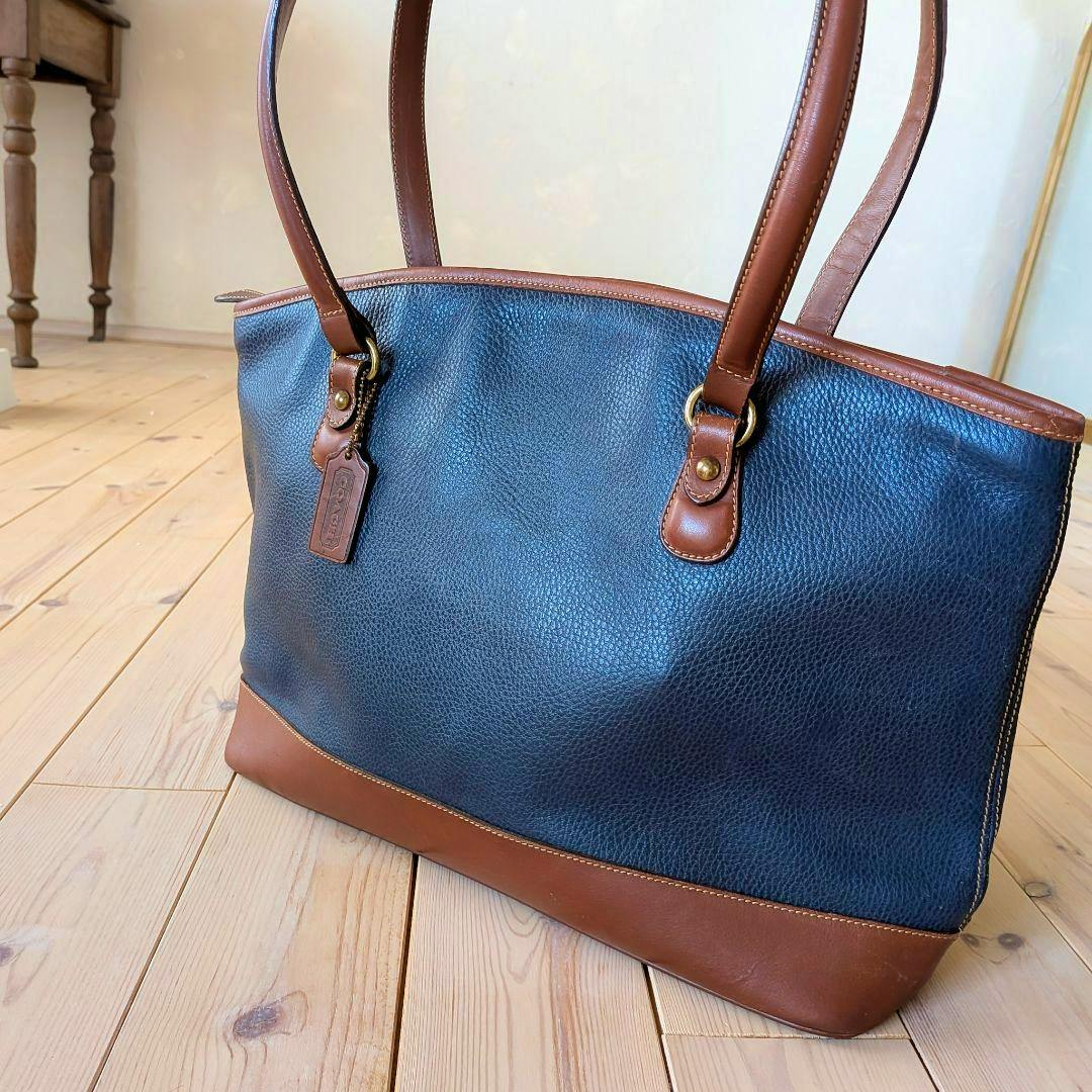 Front view of the COACH navy and brown cow leather tote bag with handles placed on a wooden floor.