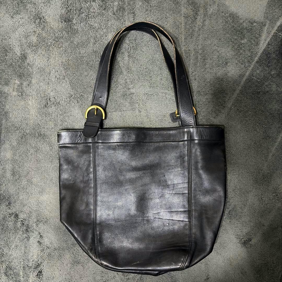 Black COACH cow leather tote bag with shoulder straps and gold-tone buckles laid flat on a gray surface.