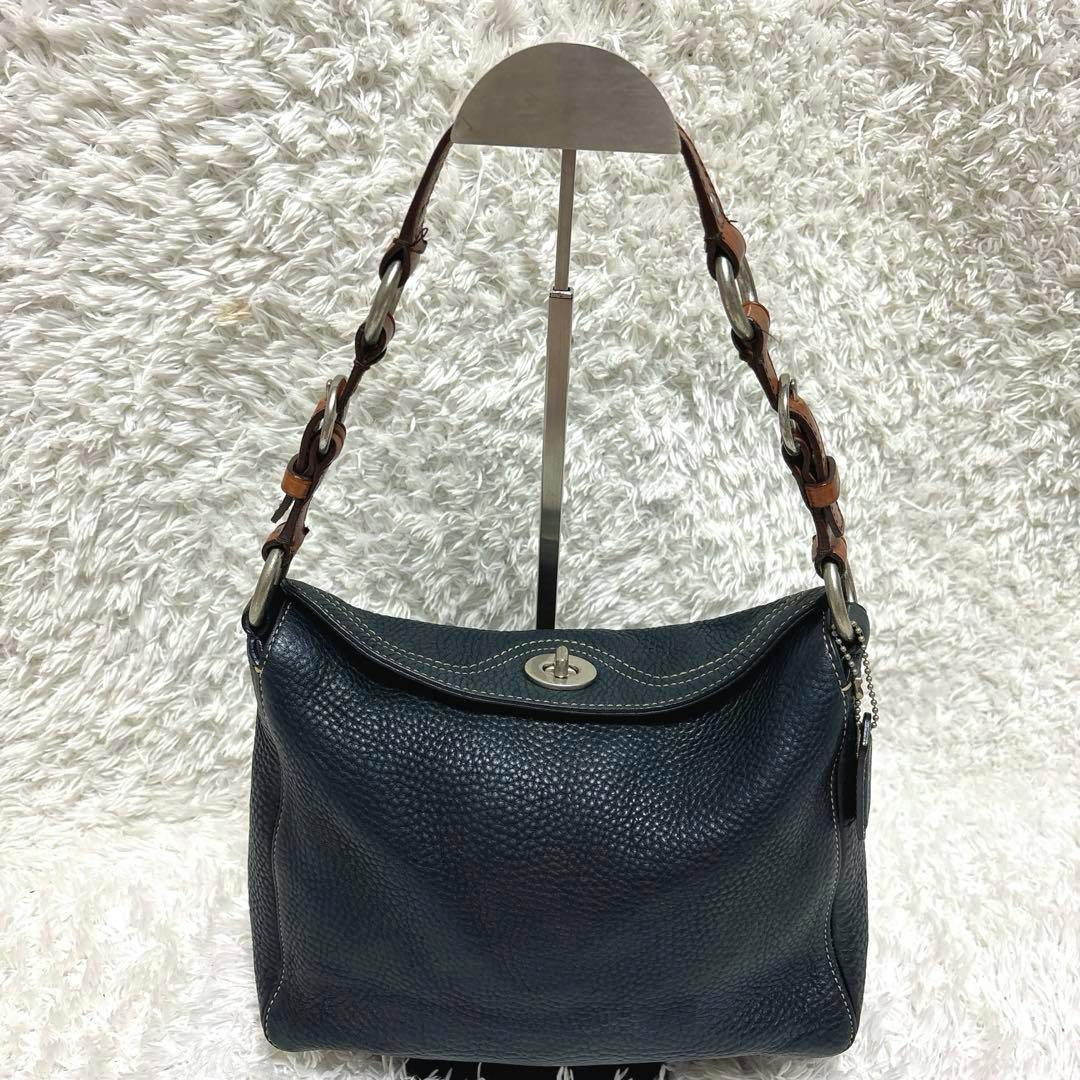 Front view of the COACH leather turn-lock one-shoulder bag on a display stand, showing the pebbled leather body, turn-lock closure, and brown strap with metal rings.