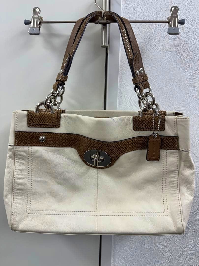 Front view of a white leather COACH tote bag with brown trim, chain-and-leather handles, and a turnlock closure hanging on a metal hanger.