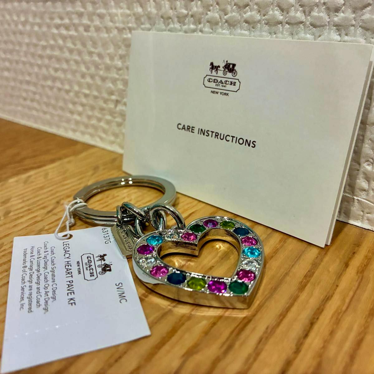 A Coach heart-shaped pave bag charm/keyring with multicolored stones attached to a metal key ring, shown with its care instructions card and product tag on a wooden surface.